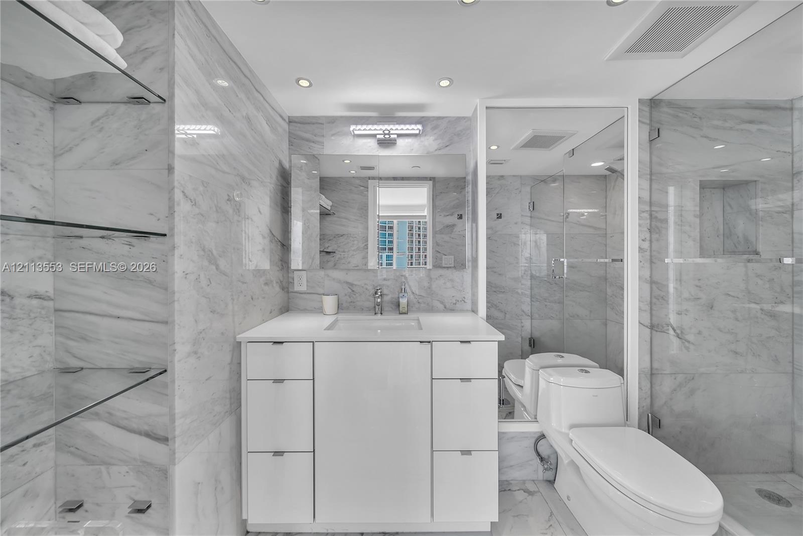 Bathroom featuring floor-to-ceiling stone-tiled walls, a white vanity with integrated basin, recessed lighting, glass shelving, and a frameless glass-enclosed shower