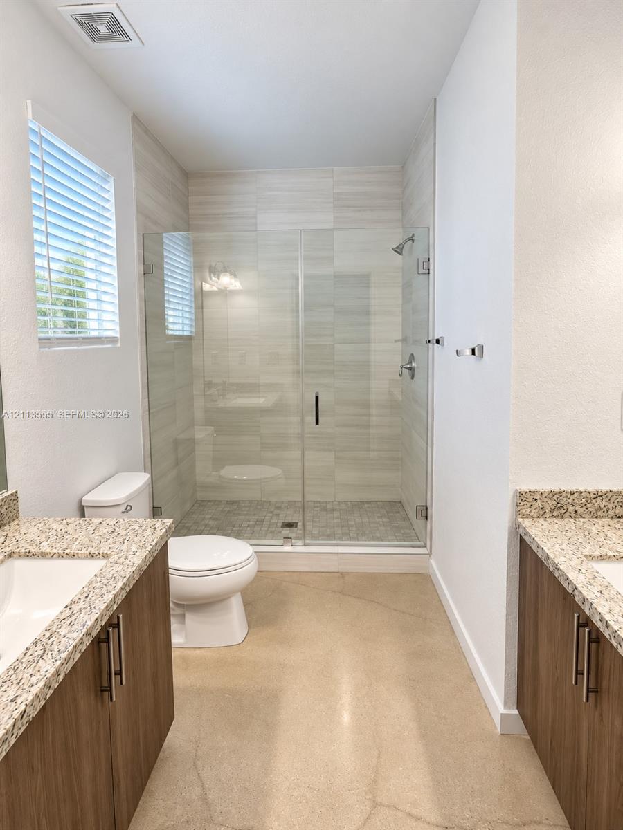 Dual wood-finish vanities with stone countertops, frameless glass shower enclosure, horizontal tile wall surround, and polished concrete flooring