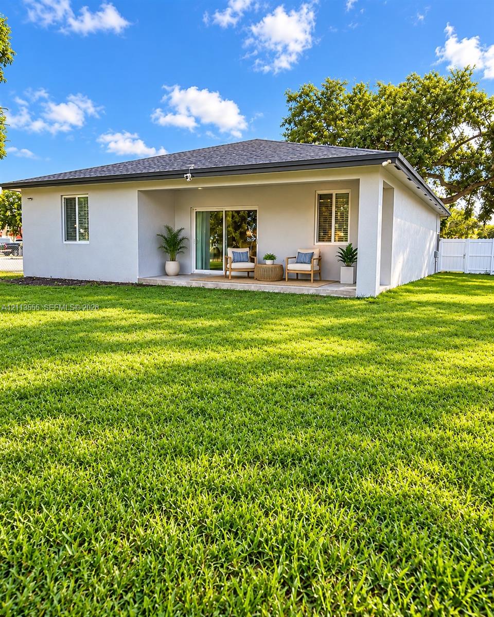 Exterior rear elevation featuring a covered concrete patio, sliding glass door, white vinyl fencing, and an expansive lawn Exterior rear elevation featuring a covered concrete patio, sliding glass door, white vinyl fencing, and an expansive lawn