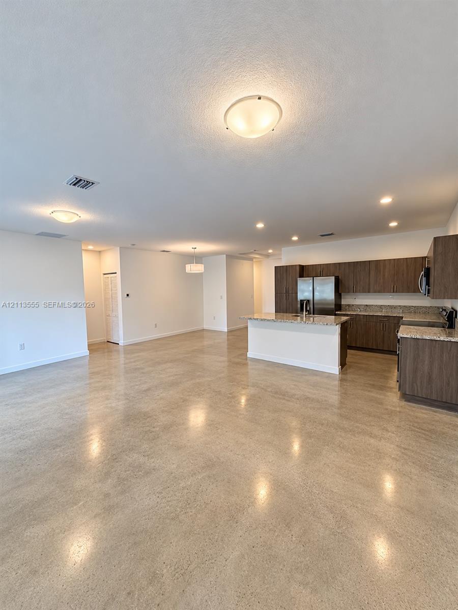 Open-concept interior featuring polished concrete flooring, kitchen with dark wood-finish cabinetry, stone-style countertops, an island, and flush-mount lighting fixtures Open-concept interior featuring polished concrete flooring, kitchen with dark wood-finish cabinetry, stone-style countertops, an island, and flush-mount lighting fixtures