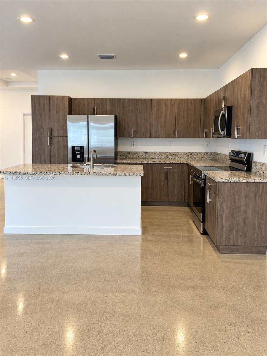 Kitchen featuring flat-panel wood-grain cabinetry, stone-finish countertops, stainless steel appliances, a center island with a double-basin sink, and polished concrete flooring
