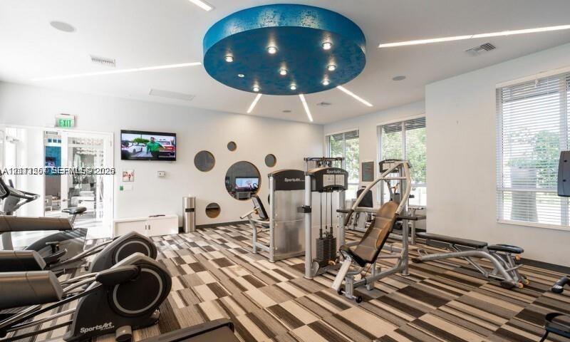 Fitness center featuring a circular recessed ceiling light feature, patterned carpet flooring, large multi-shape wall mirrors, linear recessed lighting, and expansive windows with horizontal blinds