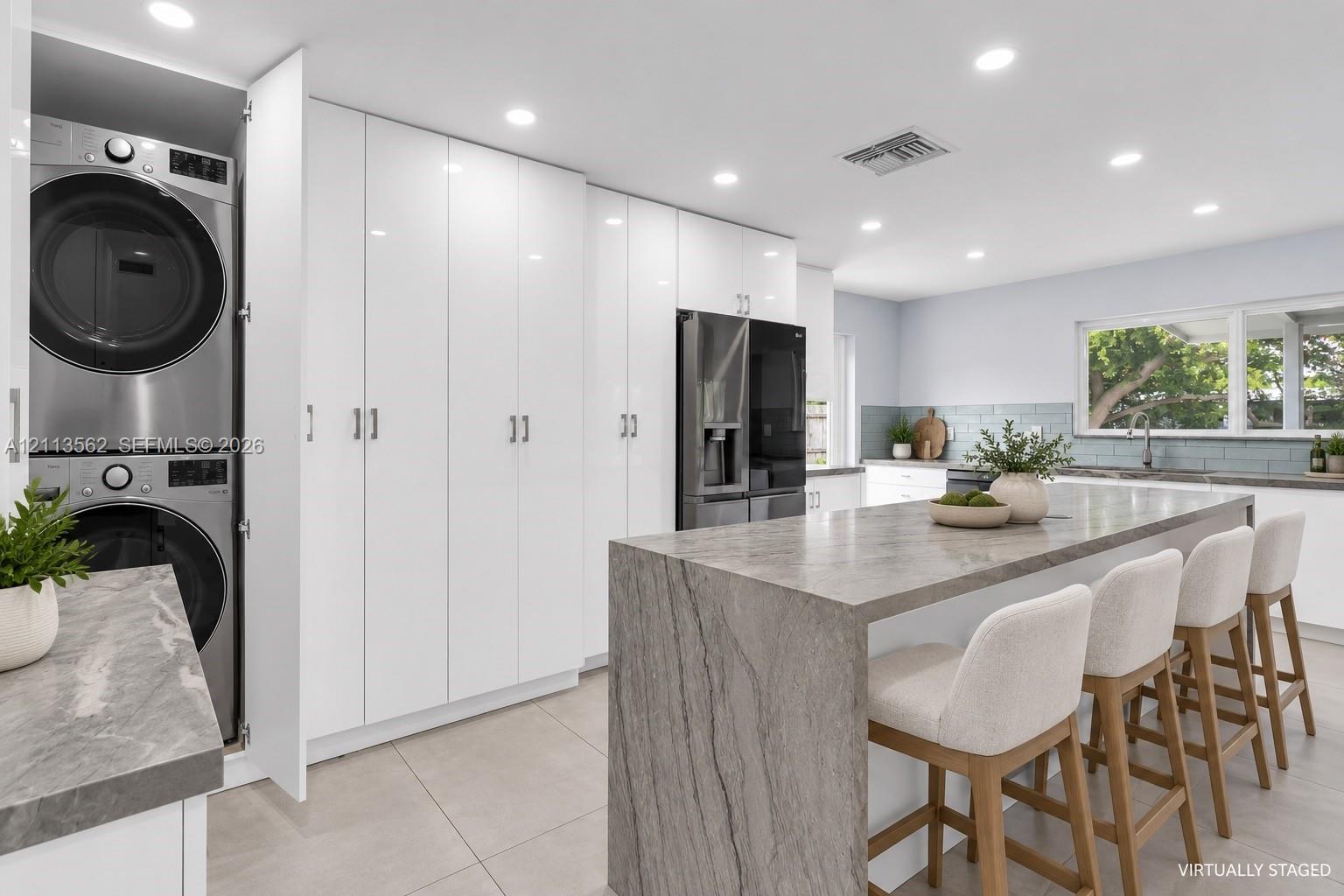 Kitchen featuring high-gloss cabinetry, a waterfall-edge island with stone-finish countertops, recessed lighting, and a concealed, built-in stacked laundry unit