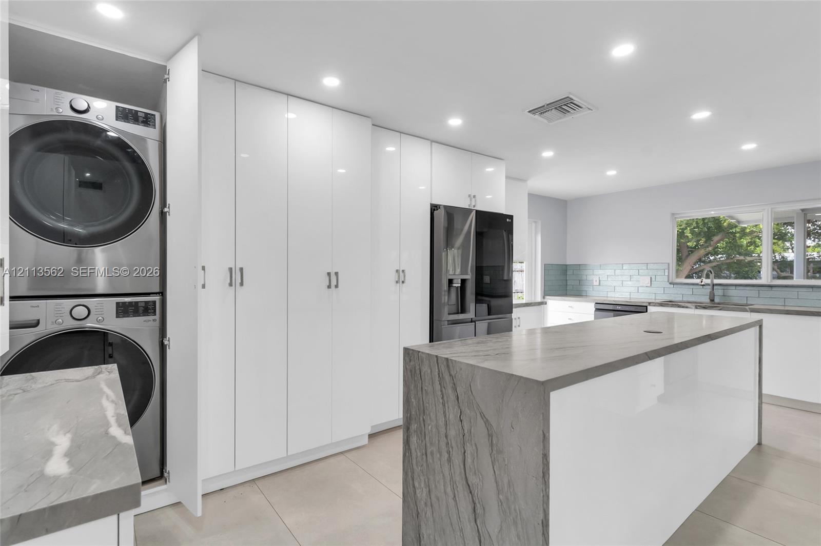 Kitchen featuring a waterfall-edge island with stone-finish countertop, high-gloss white cabinetry, integrated laundry station, and recessed lighting