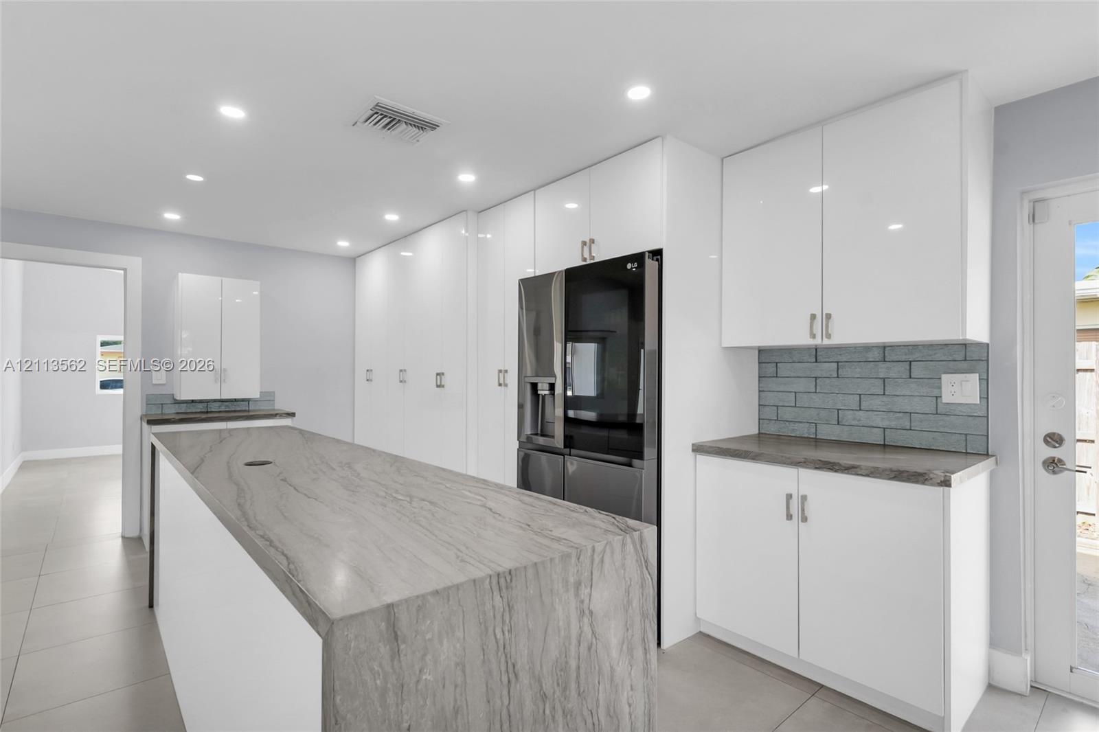 Kitchen featuring white flat-panel cabinetry, stone-surface waterfall island, grey brick-pattern backsplash, and tile flooring