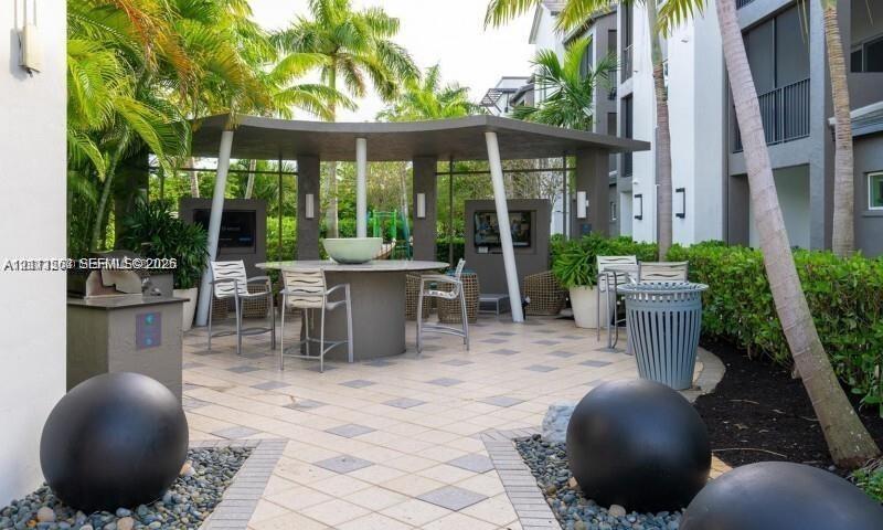 Covered outdoor pavilion with a central stone-topped table and built-in wall displays, surrounded by tile hardscaping, decorative stone spheres, and lush landscaping