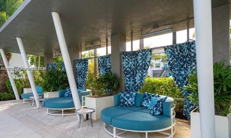 Covered outdoor lounge area featuring stone tile flooring, structural white pillars, overhead mounted lighting, and integrated planters