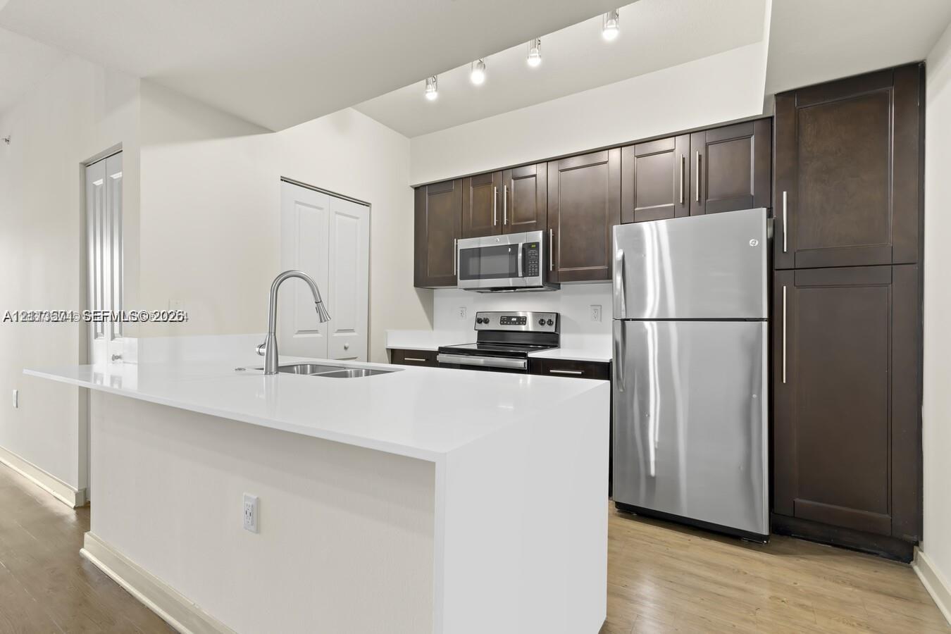 Kitchen featuring dark wood-finish cabinetry, white solid-surface countertops, a stainless steel refrigerator, a built-in microwave, and recessed track lighting