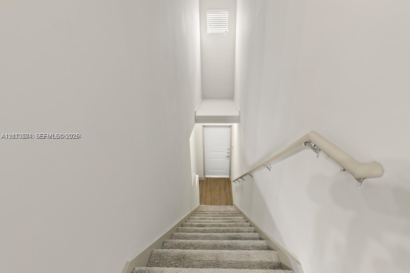 Staircase featuring neutral-toned carpeting, a wall-mounted handrail, and a direct line of sight to a white interior door with wood-finish flooring