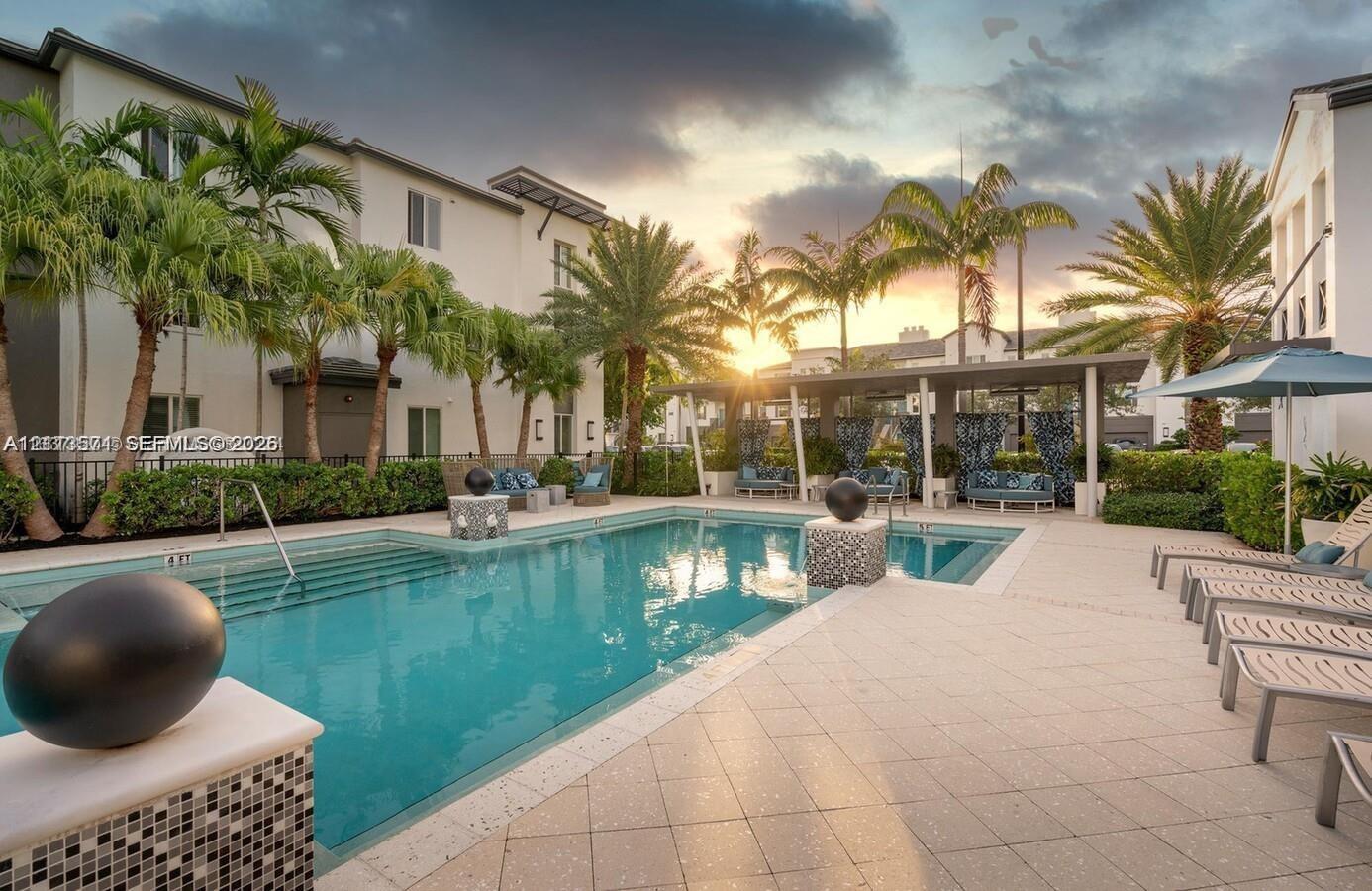 In-ground swimming pool featuring built-in entry steps, tile surround, stone-tiled decorative pillars, covered patio area, and mature palm landscaping