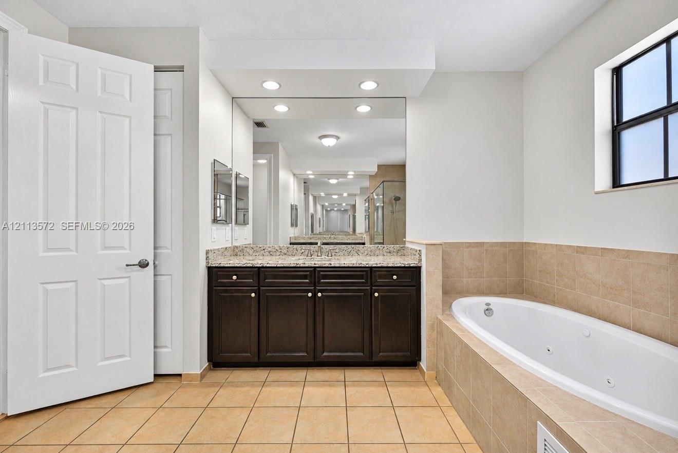 Bathroom featuring a dark-finish vanity, stone-pattern countertop, recessed lighting, wall-to-wall mirror, and a jetted soaking tub with tile surround