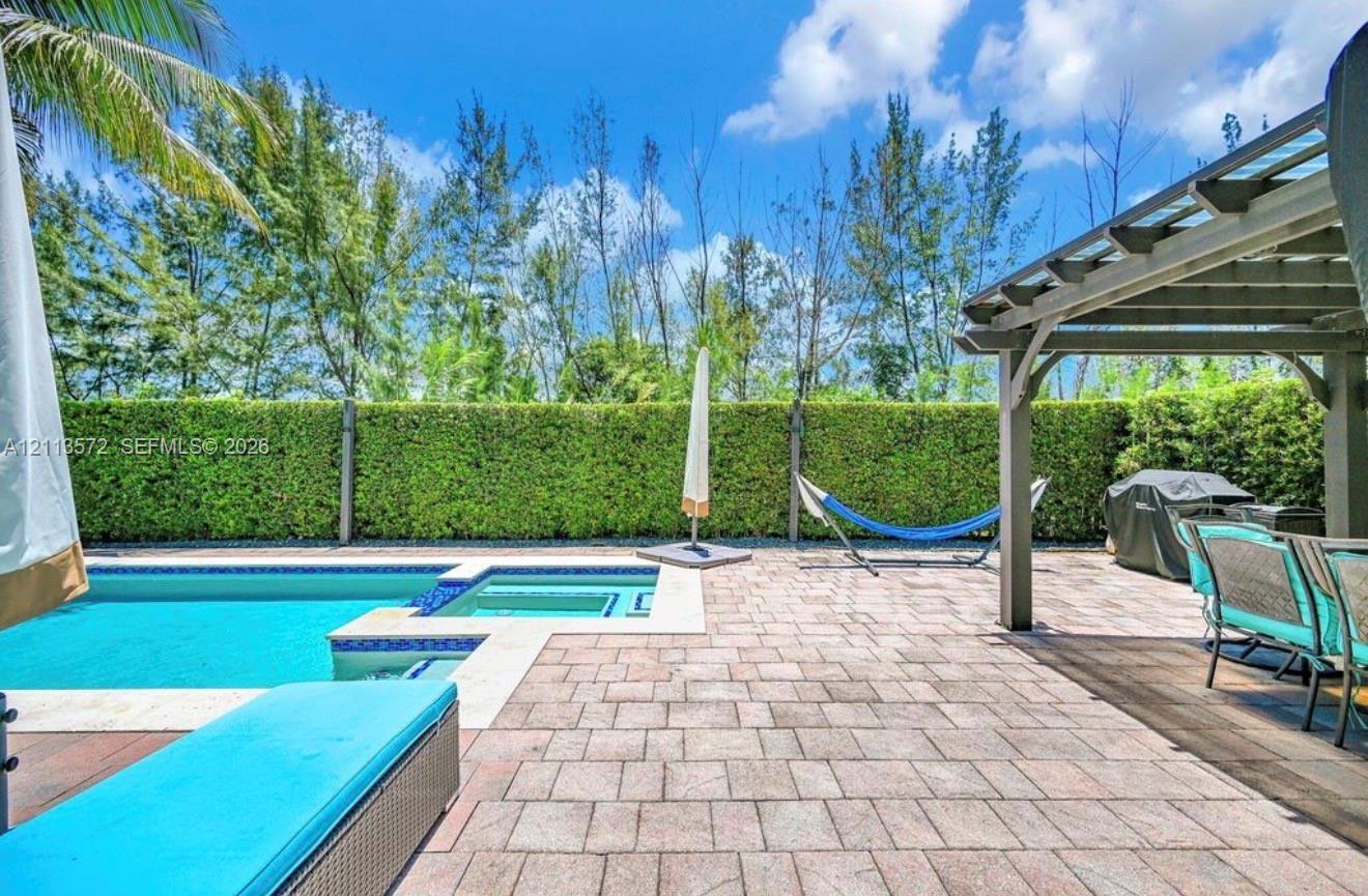 Outdoor patio area featuring a paver stone deck, a covered pergola, an in-ground swimming pool with an integrated spa, and manicured hedge fencing