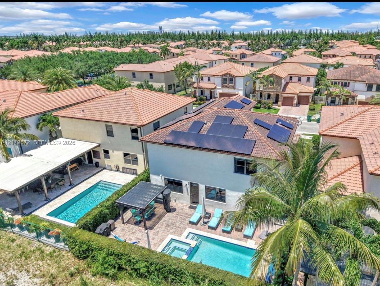 Aerial perspective featuring a two-story residence equipped with roof-mounted solar panels, a rectangular swimming pool, a spillover spa, a stone-paved patio, and a covered lanai