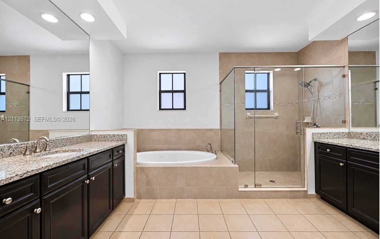 Dual vanities with stone countertops and dark cabinetry, large soaking tub with tile surround, and walk-in glass-enclosed shower with tiled walls