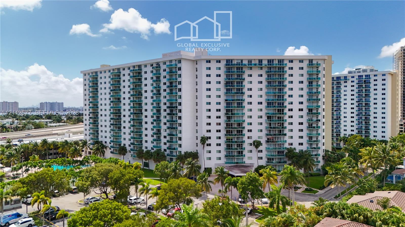 High-rise residential building featuring stacked private balconies with glass railings, an exterior swimming pool, mature palm tree landscaping, and paved parking areas High-rise residential building featuring stacked private balconies with glass railings, an exterior swimming pool, mature palm tree landscaping, and paved parking areas
