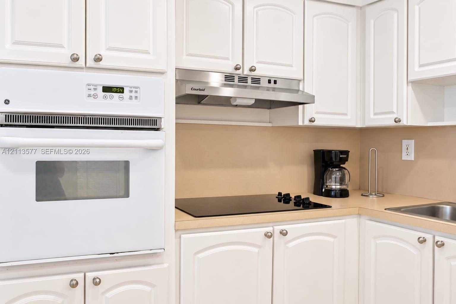 Kitchen featuring white raised-panel cabinetry, built-in white wall oven, stainless steel range hood, smooth-top electric cooktop, and solid-surface countertops Kitchen featuring white raised-panel cabinetry, built-in white wall oven, stainless steel range hood, smooth-top electric cooktop, and solid-surface countertops