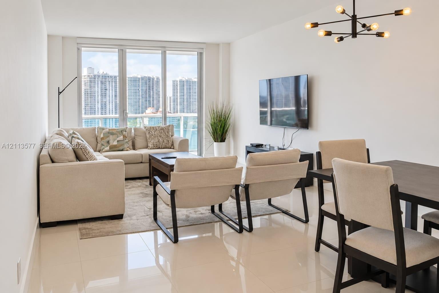 Living area featuring large-format tile flooring, floor-to-ceiling windows with a glass balcony railing, and a contemporary multi-arm chandelier Living area featuring large-format tile flooring, floor-to-ceiling windows with a glass balcony railing, and a contemporary multi-arm chandelier