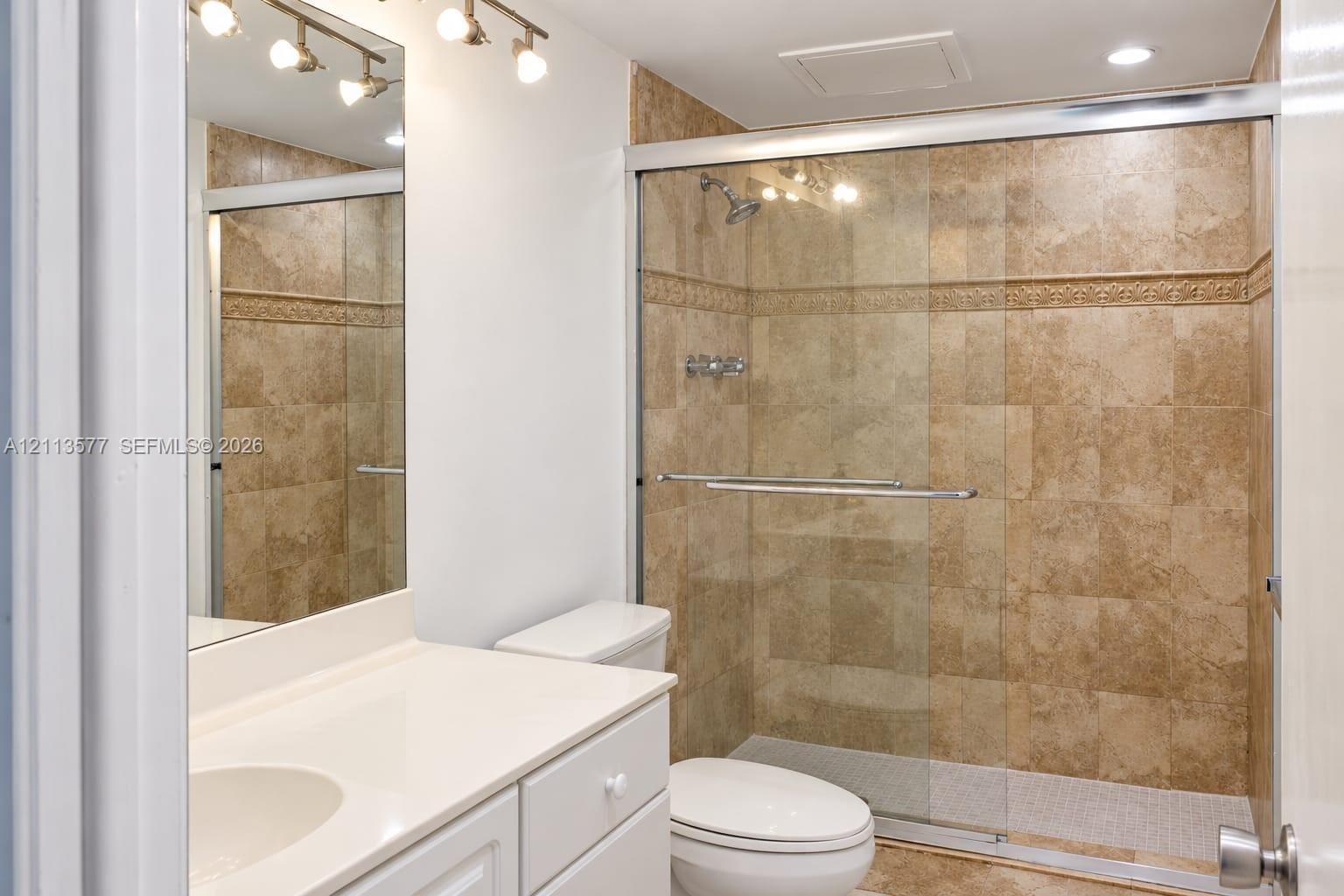 Bathroom featuring a vanity with a white countertop, an integrated sink, a full-length wall mirror, multi-light fixture, and a shower enclosure with floor-to-ceiling stone-finish tile surround and sliding glass doors