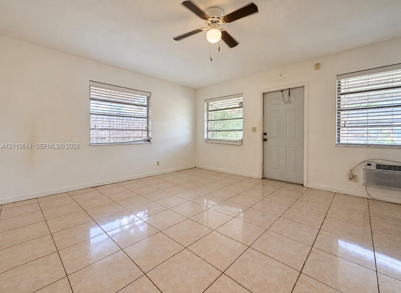 Neutral-toned tile flooring, three windows with horizontal blinds, a ceiling fan with integrated lighting, and a wall-mounted air conditioning unit