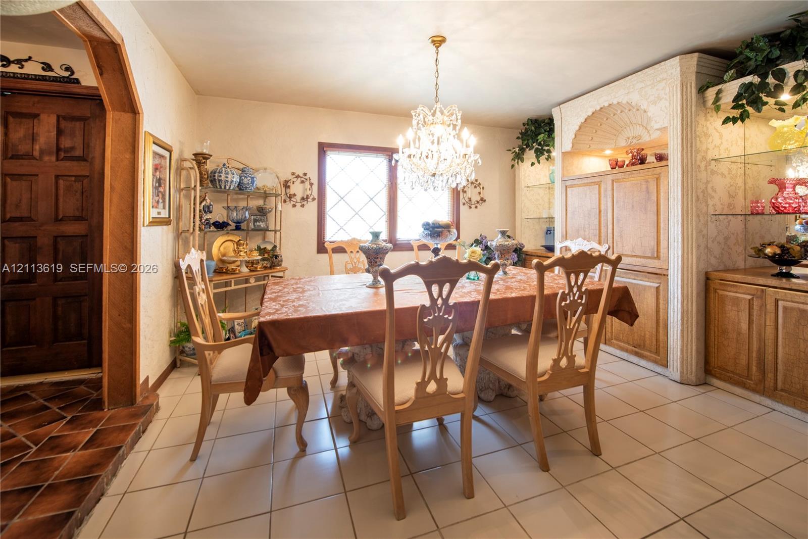 Dining area featuring tiled flooring, a crystal chandelier, arched wooden doorway, and built-in cabinetry with glass shelving Dining area featuring tiled flooring, a crystal chandelier, arched wooden doorway, and built-in cabinetry with glass shelving