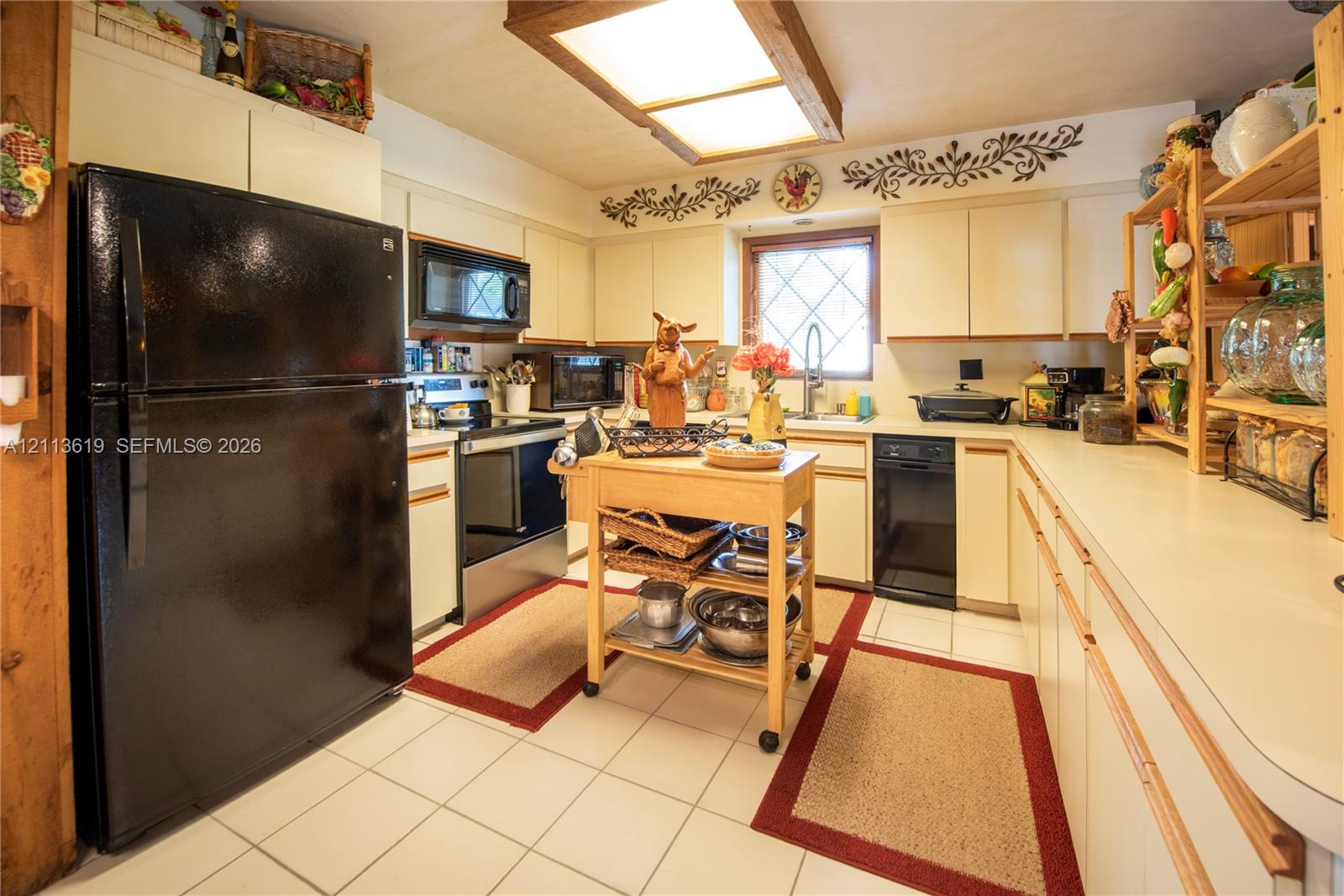 Kitchen featuring flat-panel cabinetry, a black refrigerator, a black dishwasher, a recessed ceiling light fixture with wood-trim, and ceramic tile flooring Kitchen featuring flat-panel cabinetry, a black refrigerator, a black dishwasher, a recessed ceiling light fixture with wood-trim, and ceramic tile flooring