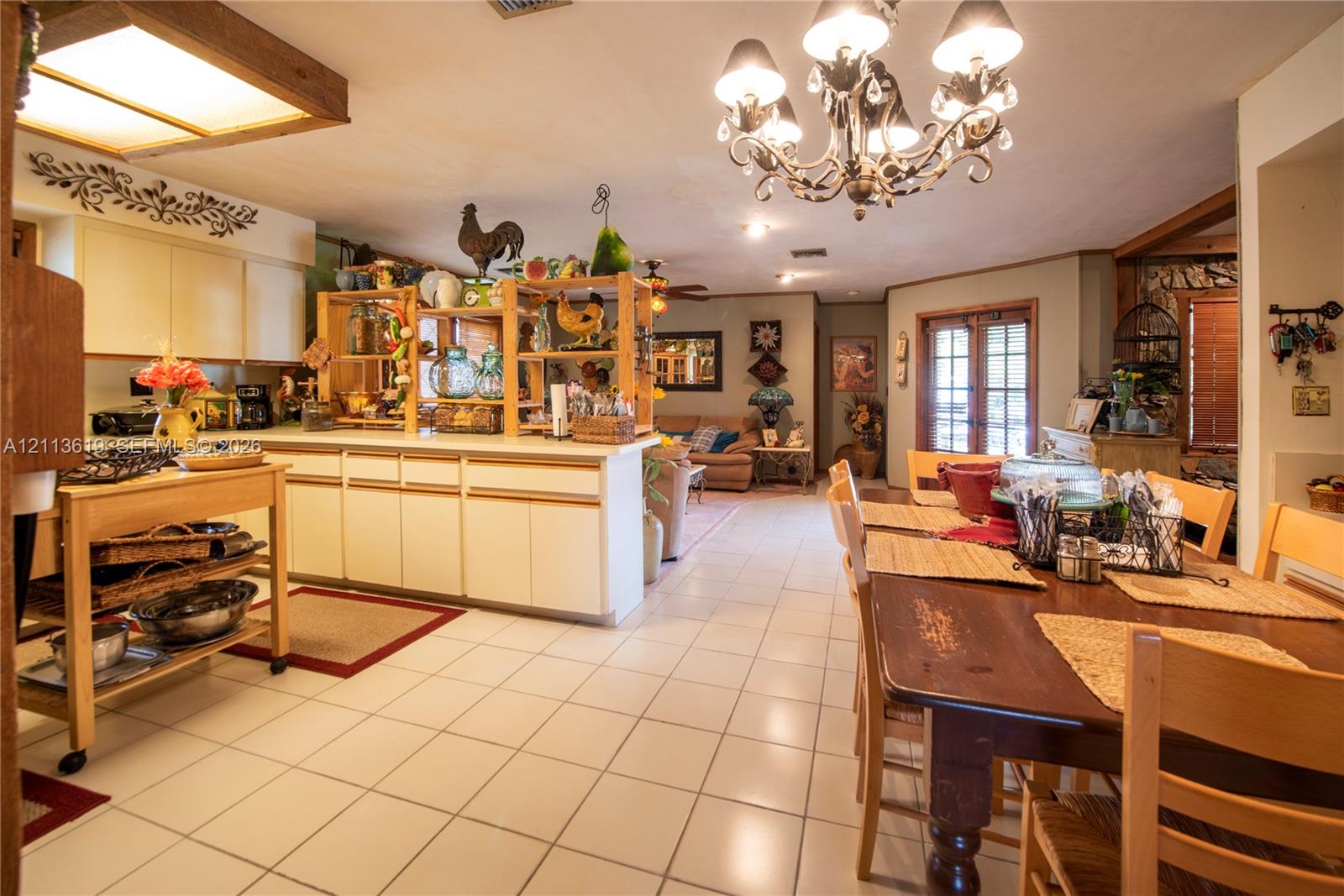 Open-concept kitchen featuring light-toned cabinetry, ceramic tile flooring, a multi-arm chandelier with decorative glass elements, and double-door access to exterior space Open-concept kitchen featuring light-toned cabinetry, ceramic tile flooring, a multi-arm chandelier with decorative glass elements, and double-door access to exterior space