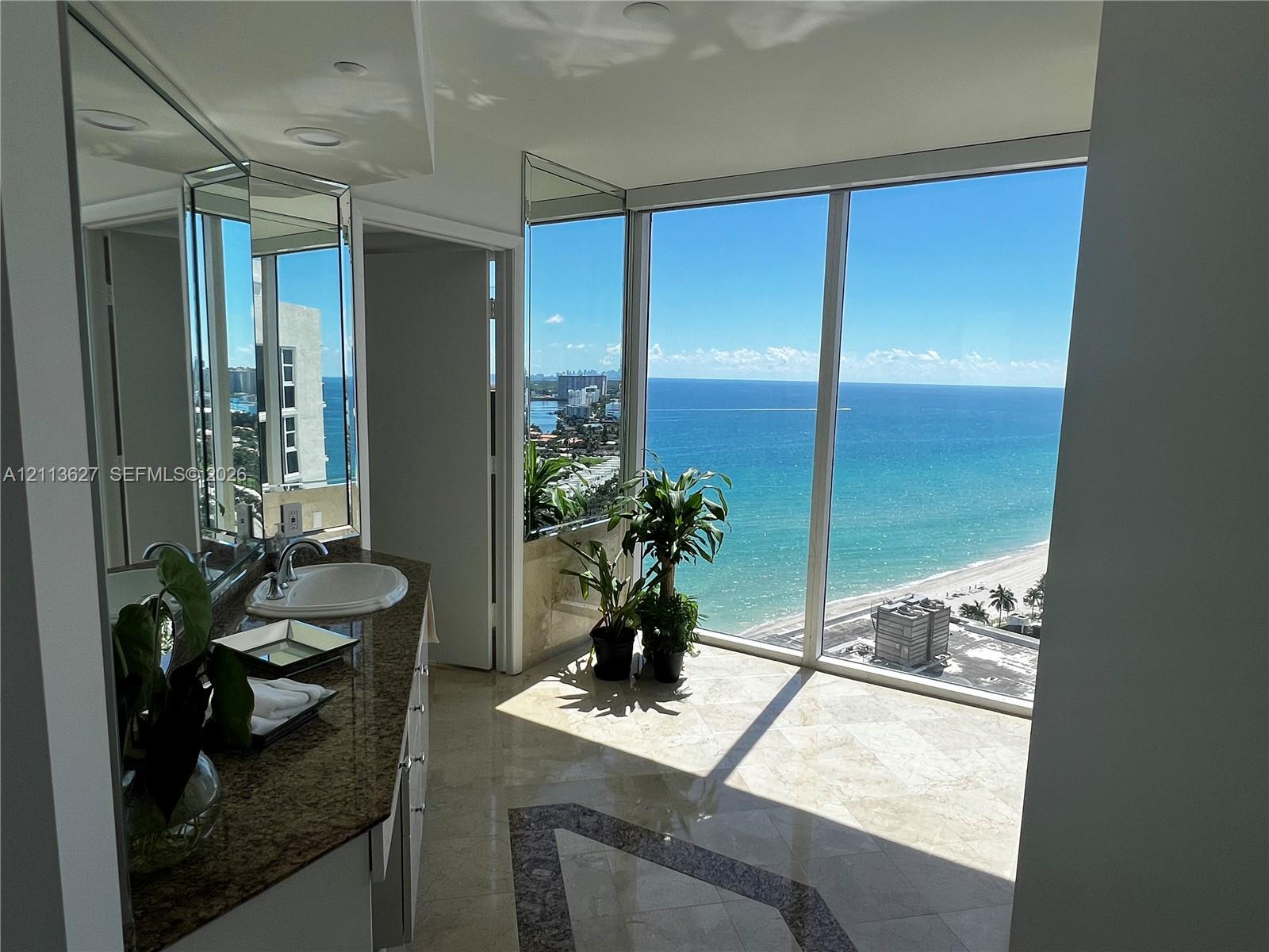 Floor-to-ceiling windows, stone tile flooring with decorative inlay, expansive vanity with integrated sink, and full-wall mirrors overlooking the ocean coastline