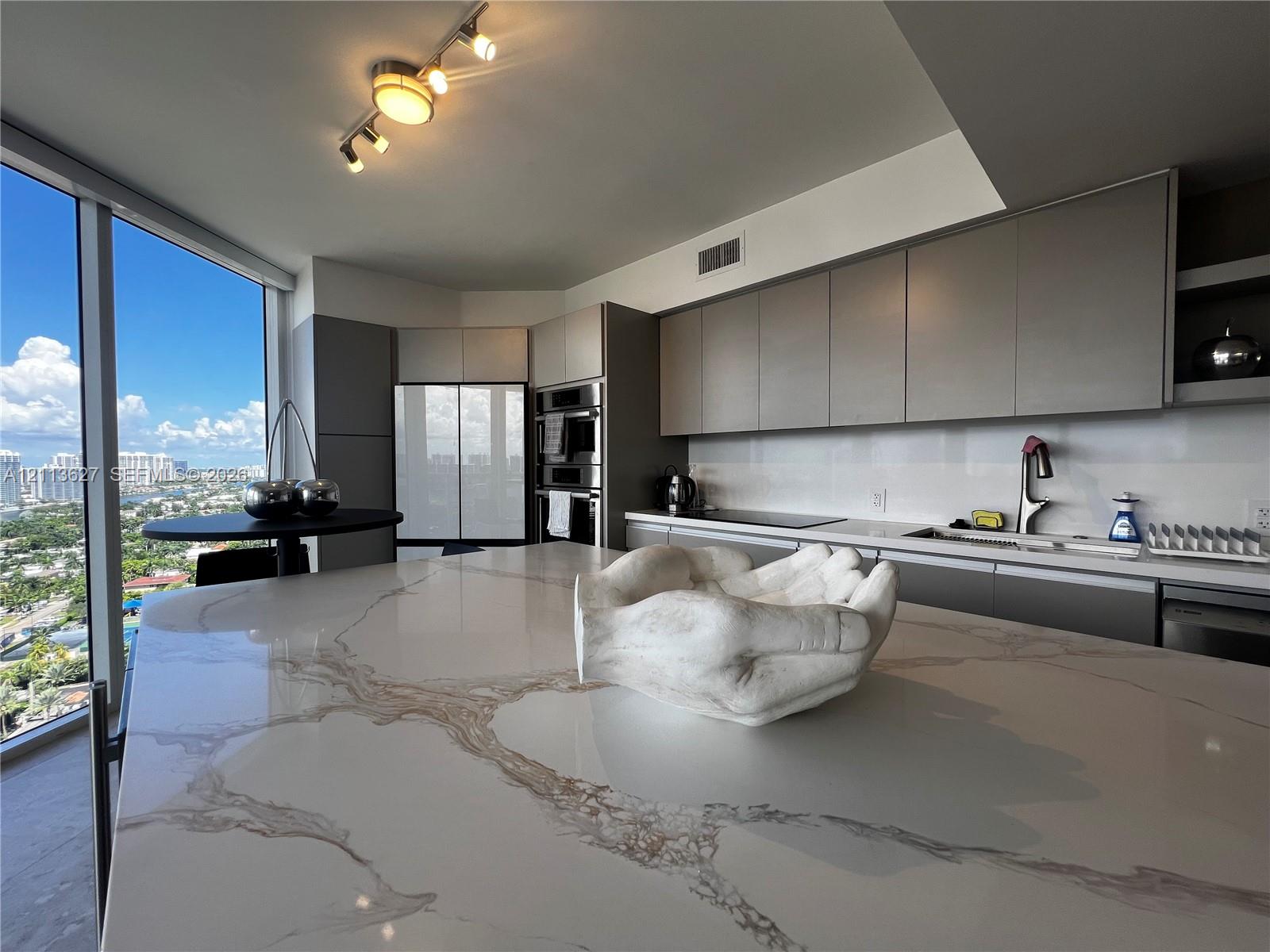 Kitchen featuring an oversized island with a stone-finish countertop, flat-panel cabinetry, integrated wall ovens, and floor-to-ceiling windows