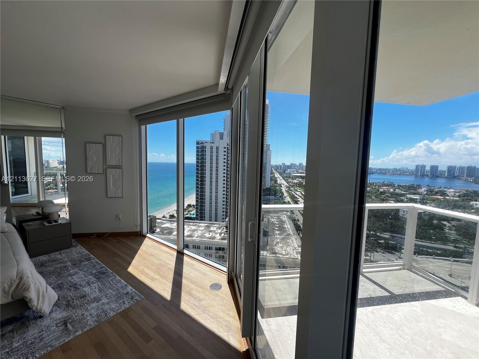 Floor-to-ceiling glass windows, expansive private balcony with glass railing, wood-finish flooring, and direct coastal and city panoramas