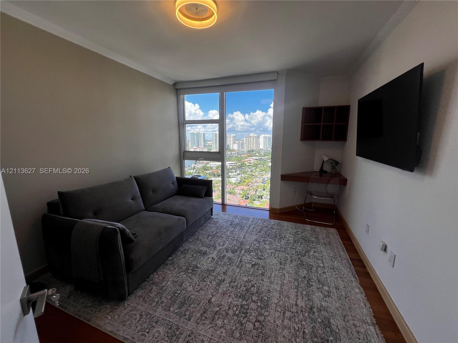 Floor-to-ceiling windows featuring urban scenery, built-in corner desk with integrated shelving, wood-finish flooring, and circular flush-mount ceiling lighting