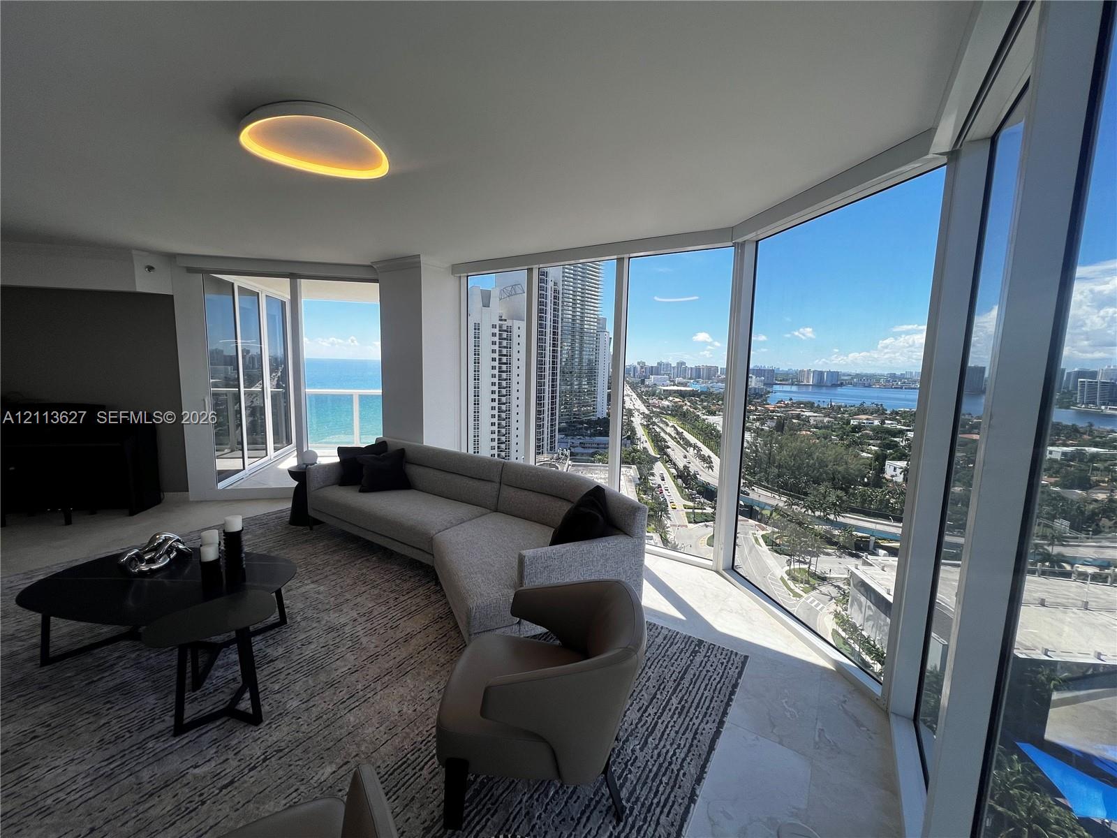 Floor-to-ceiling glass walls, wrap-around windows, stone flooring, minimalist circular ceiling fixture, and a glass-railed balcony