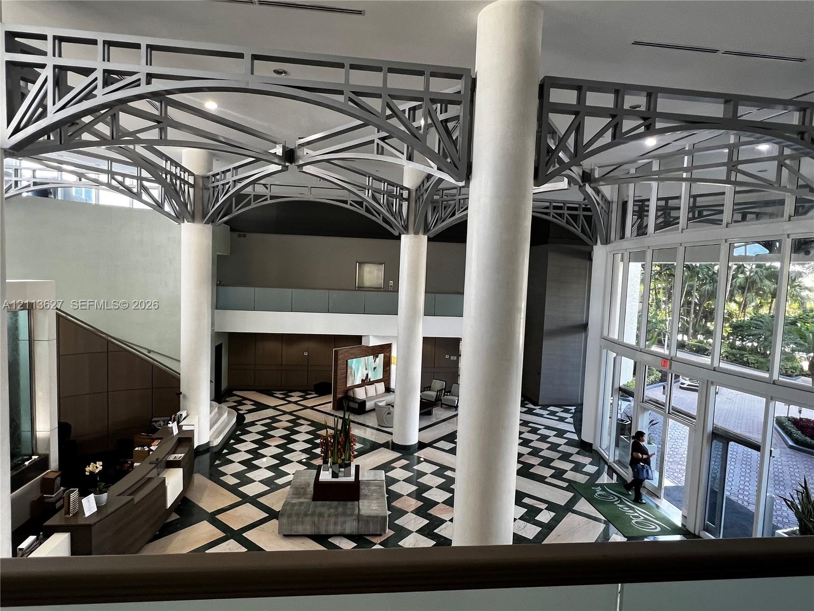 Grand foyer featuring vaulted structural steel ceiling trusses, floor-to-ceiling windows, polished stone flooring in a geometric pattern, and prominent cylindrical columns