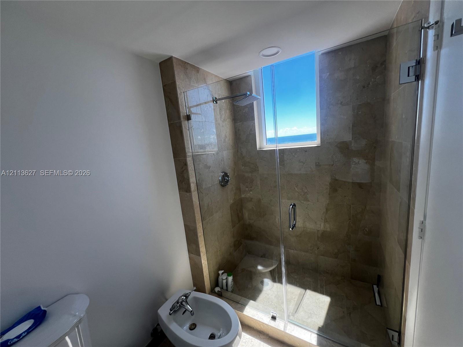 Walk-in shower featuring tile surround, glass enclosure, and built-in bench, complemented by a bidet and natural light from an integrated window