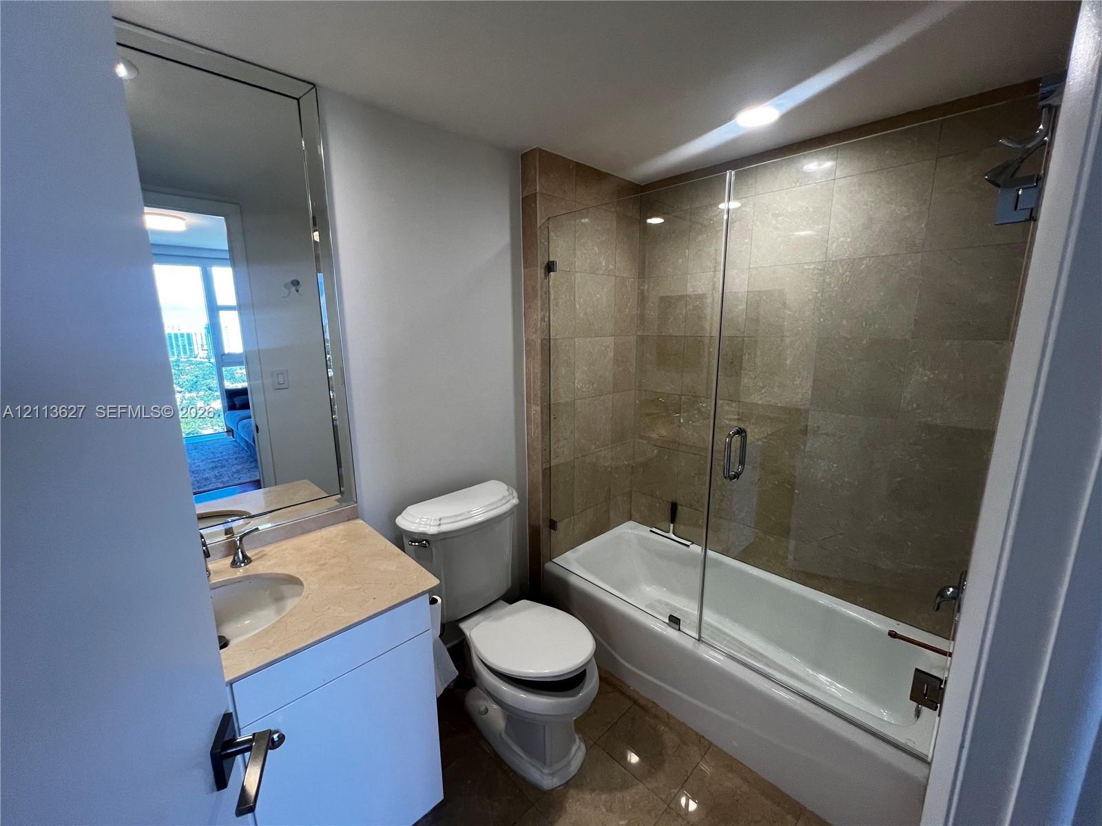 Bathroom featuring a stone-topped vanity with an undermount sink, a large wall-mounted mirror, and a tile-surround tub with a glass panel enclosure