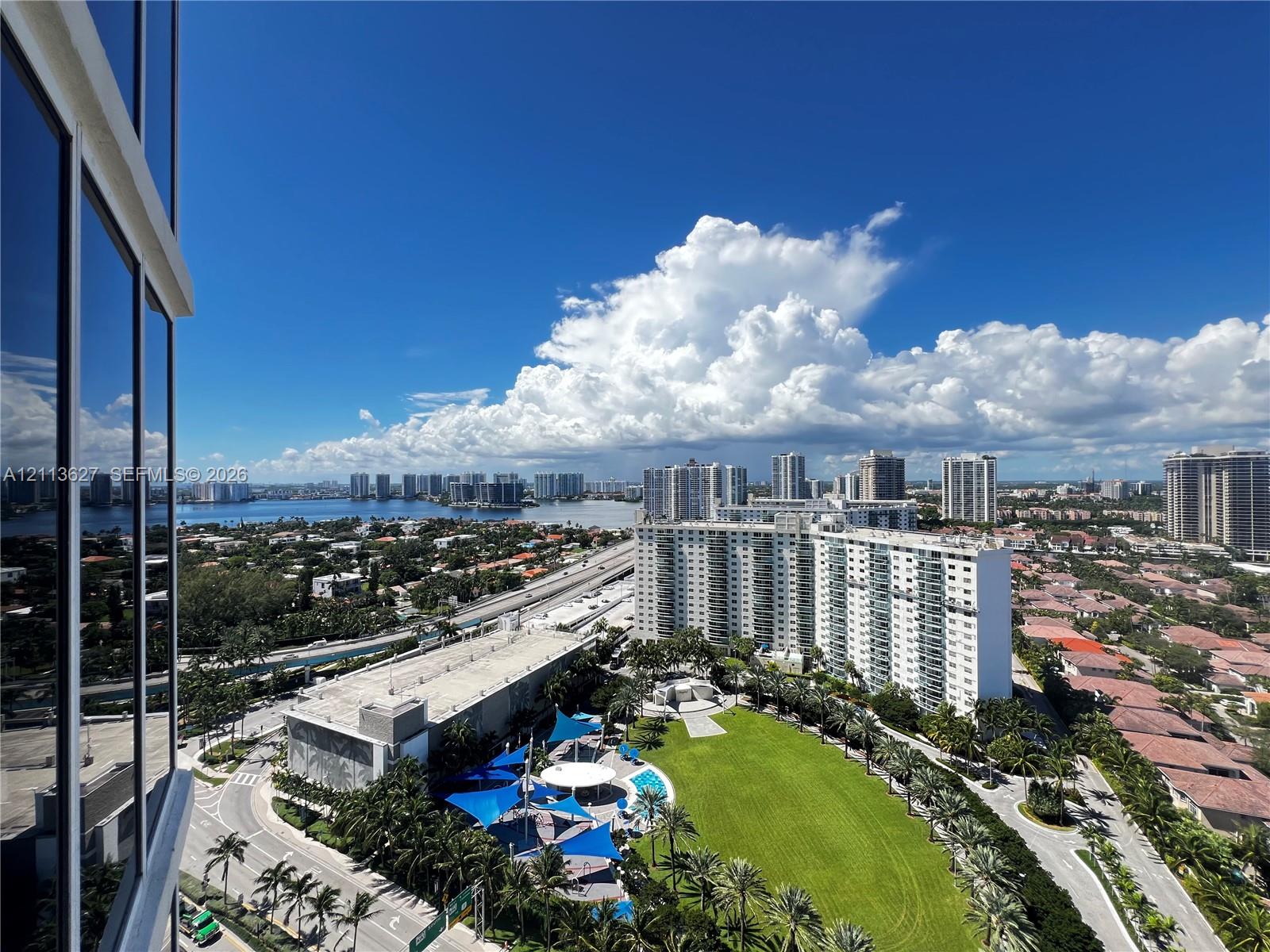 High-rise perspective featuring glass curtain wall facade, expansive green space with mature palms, and structured recreation area with shade sails