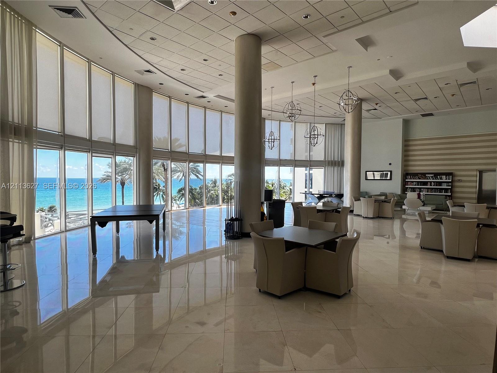 Expansive common area featuring floor-to-ceiling windows, polished stone flooring, structural columns, and spherical pendant lighting, with direct exposure to the coast