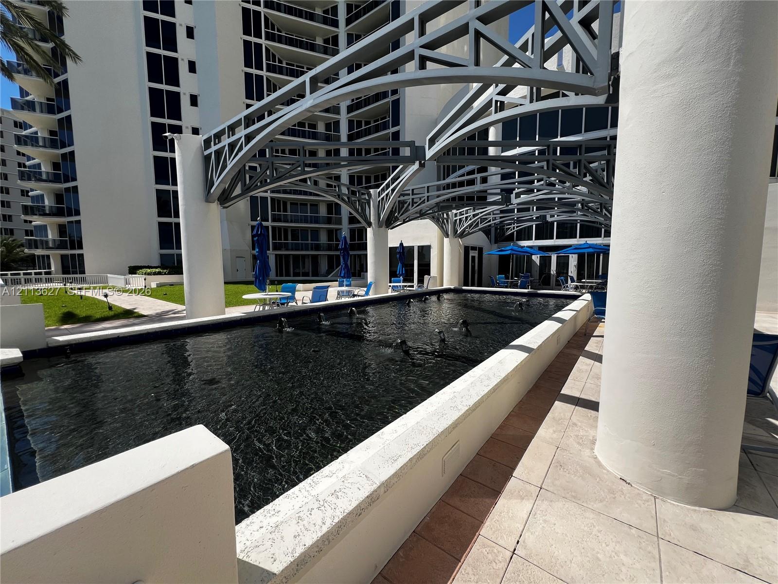 Rectangular reflective pool with built-in water features, architectural overhead metal framing, cylindrical support columns, and light-toned stone tile paving