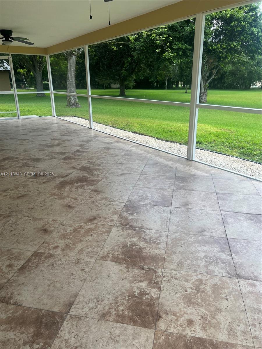 Screened-in patio featuring tile flooring, a ceiling fan, and a gravel border overlooking a grass lawn with mature trees