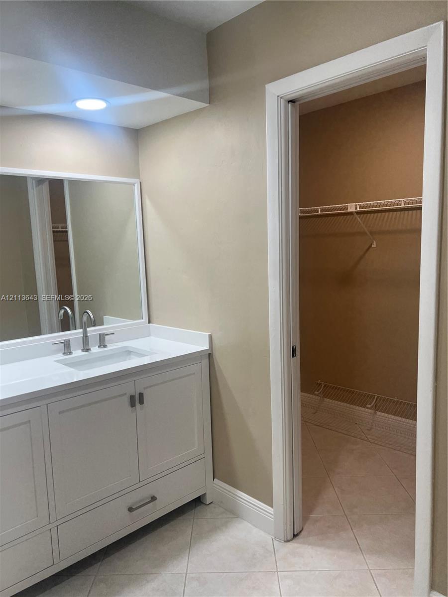 White vanity cabinet featuring a rectangular undermount sink, stone-finish countertop, and brushed-nickel hardware, accompanied by a framed mirror, recessed lighting, and a walk-in closet with built-in wire shelving