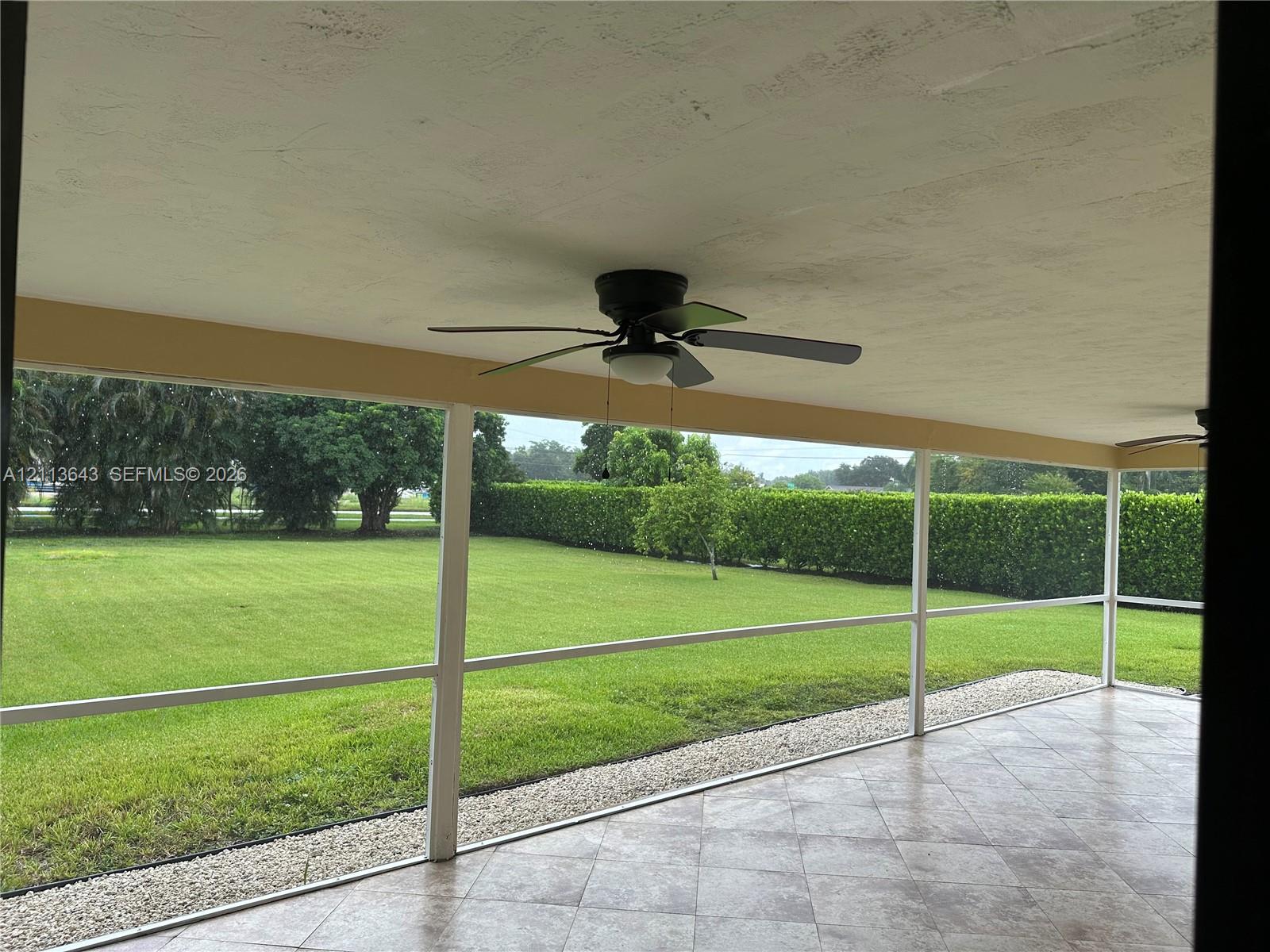 Screened-in patio featuring tile flooring, a ceiling fan, and access to a level lawn with established hedging