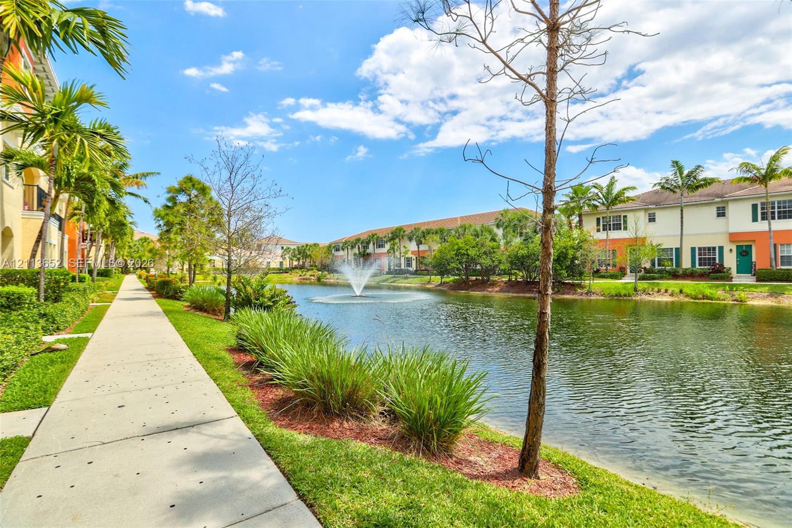 Community grounds featuring a central pond, a fountain, a paved walking path, mature palm trees, and manicured landscaping