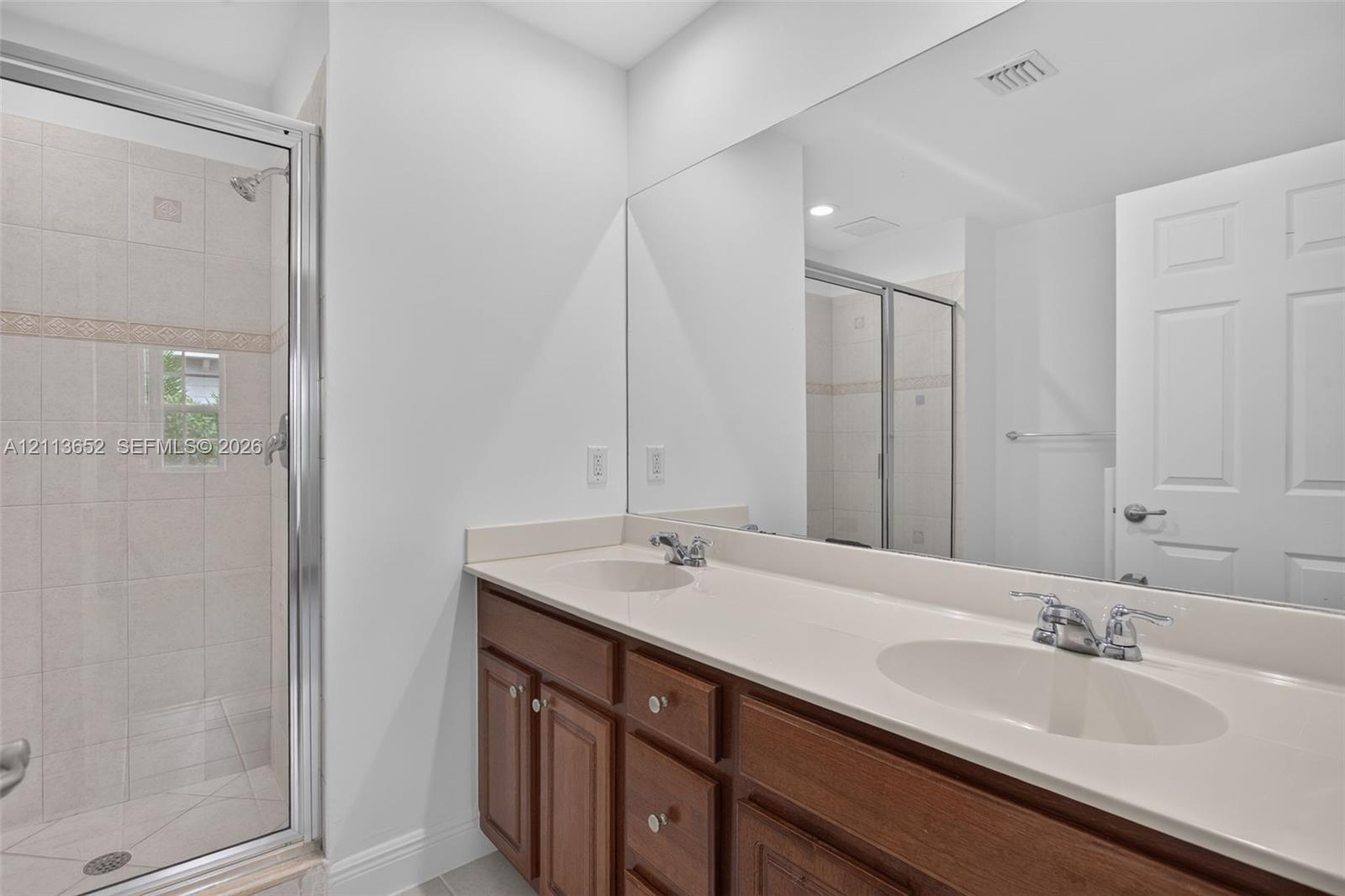 Double vanity with integrated sinks, neutral countertop, wood cabinetry, and a full-width wall mirror, alongside a walk-in shower with tiled surround and glass enclosure