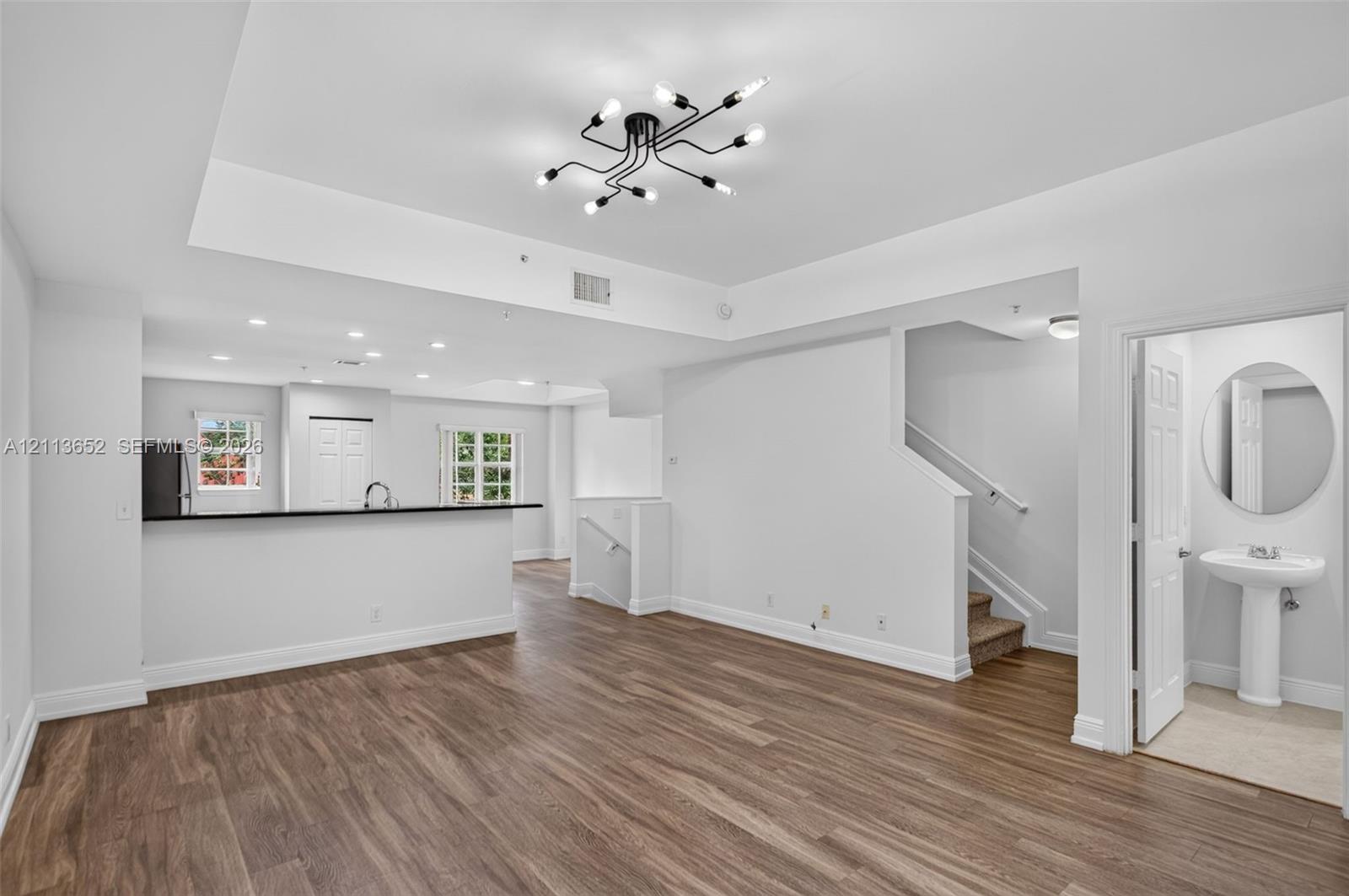 Open-concept interior featuring wood-finish flooring, a multi-arm modern chandelier, tray ceilings, and a powder room with a pedestal sink and circular mirror