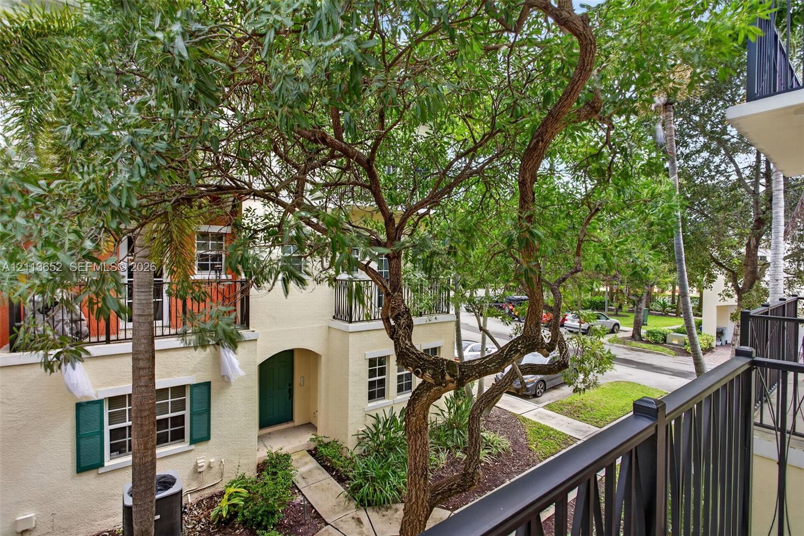 Stucco exterior with decorative green shutters, wrought iron balcony railings, arched entryway, and mature landscaping