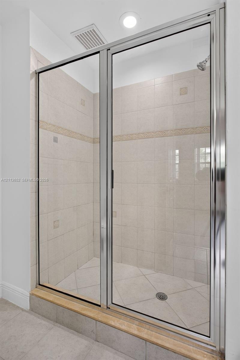 Walk-in shower enclosure featuring a polished metal frame, light-toned ceramic wall tiling with a decorative border inlay, diamond-patterned floor tile, and a raised stone threshold