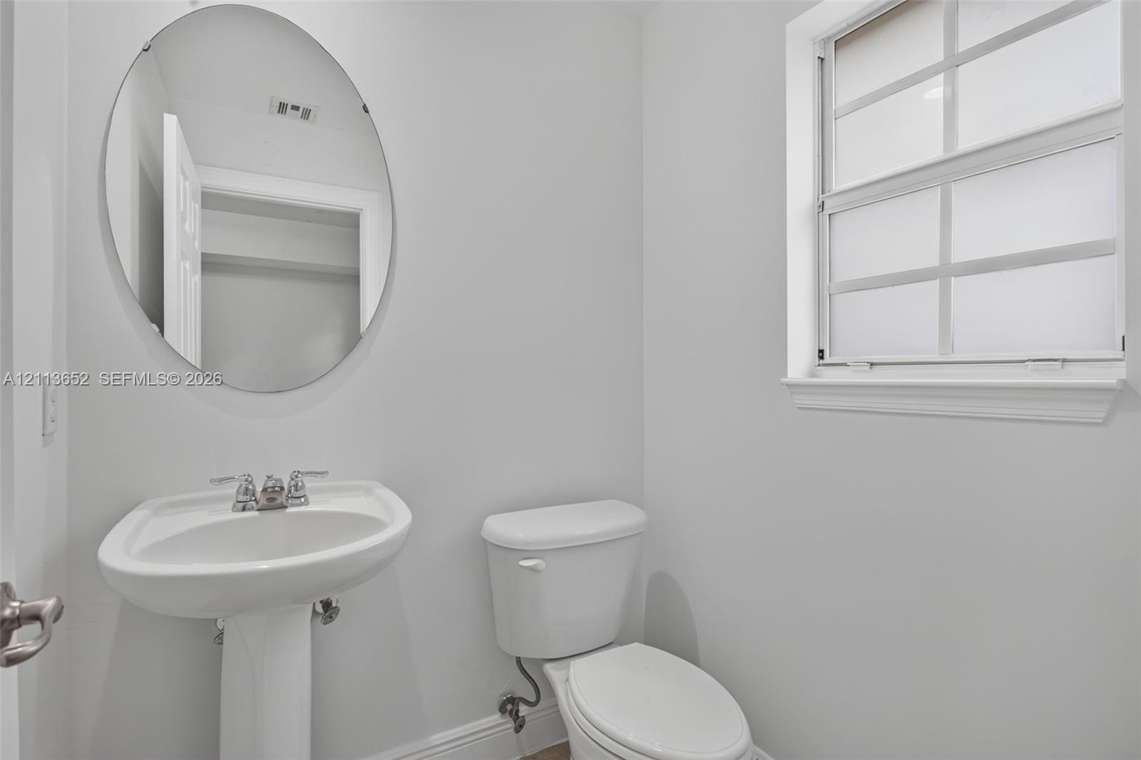 White pedestal sink with chrome faucet, oval frameless mirror, and multi-pane window with interior muntins