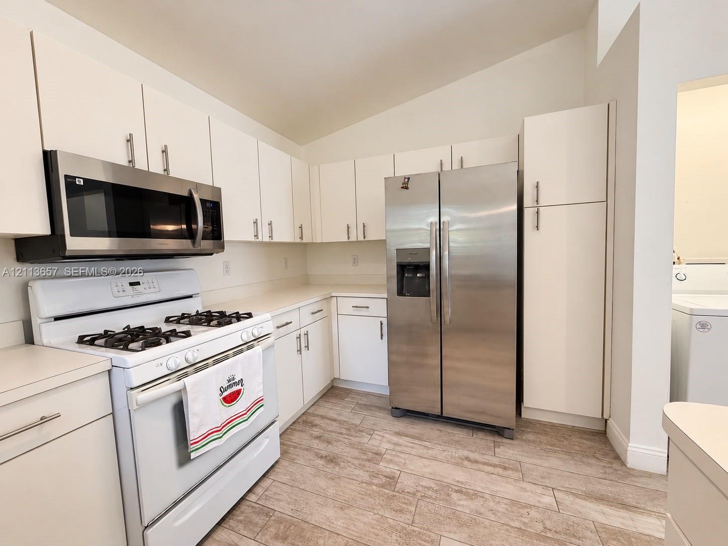 Kitchen featuring white cabinetry, wood-finish flooring, vaulted ceiling, and stainless steel appliances including a built-in microwave and side-by-side refrigerator Kitchen featuring white cabinetry, wood-finish flooring, vaulted ceiling, and stainless steel appliances including a built-in microwave and side-by-side refrigerator