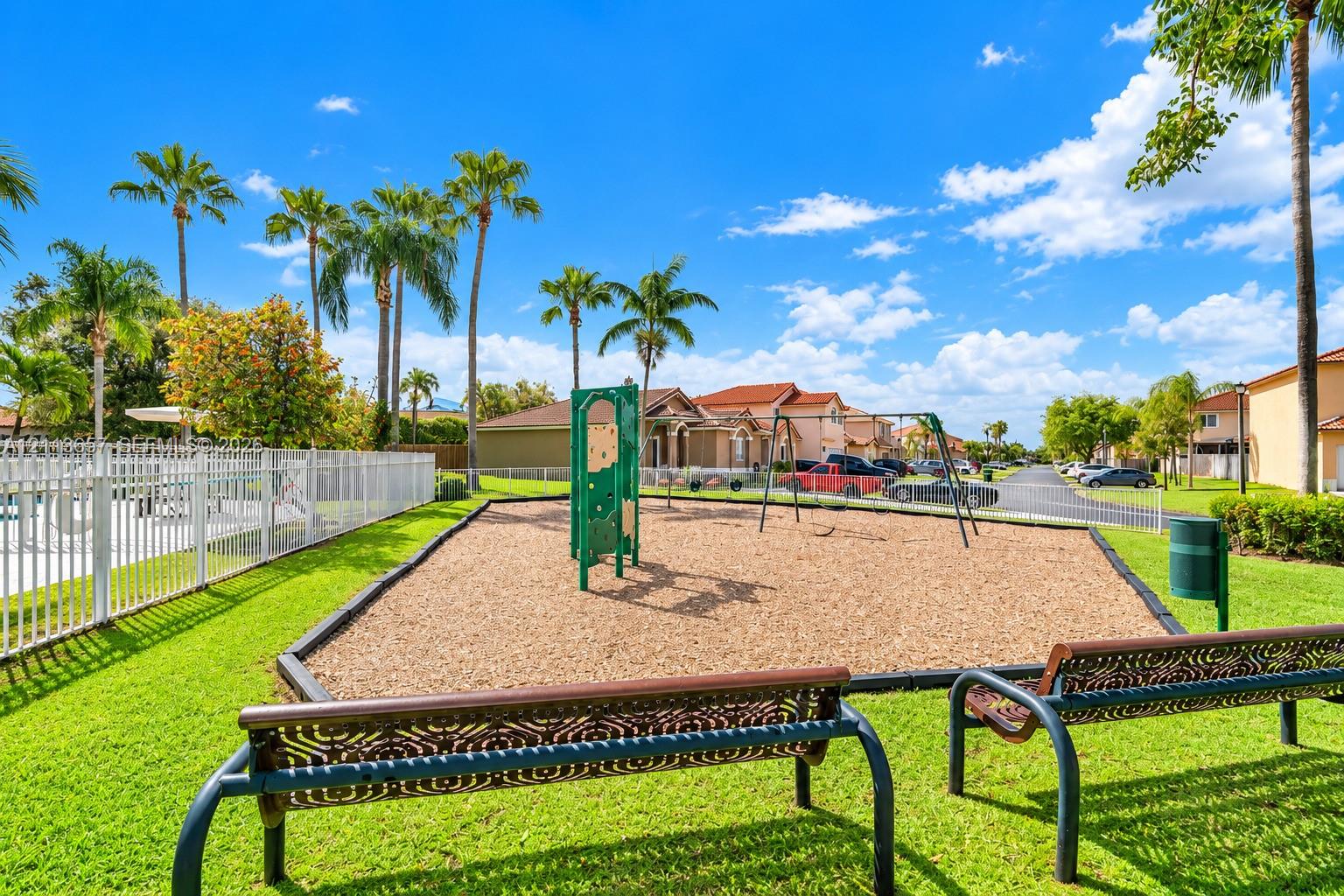Community playground featuring a climbing structure, swing set, wood-chip ground cover, and metal bench seating surrounded by manicured turf and palm trees