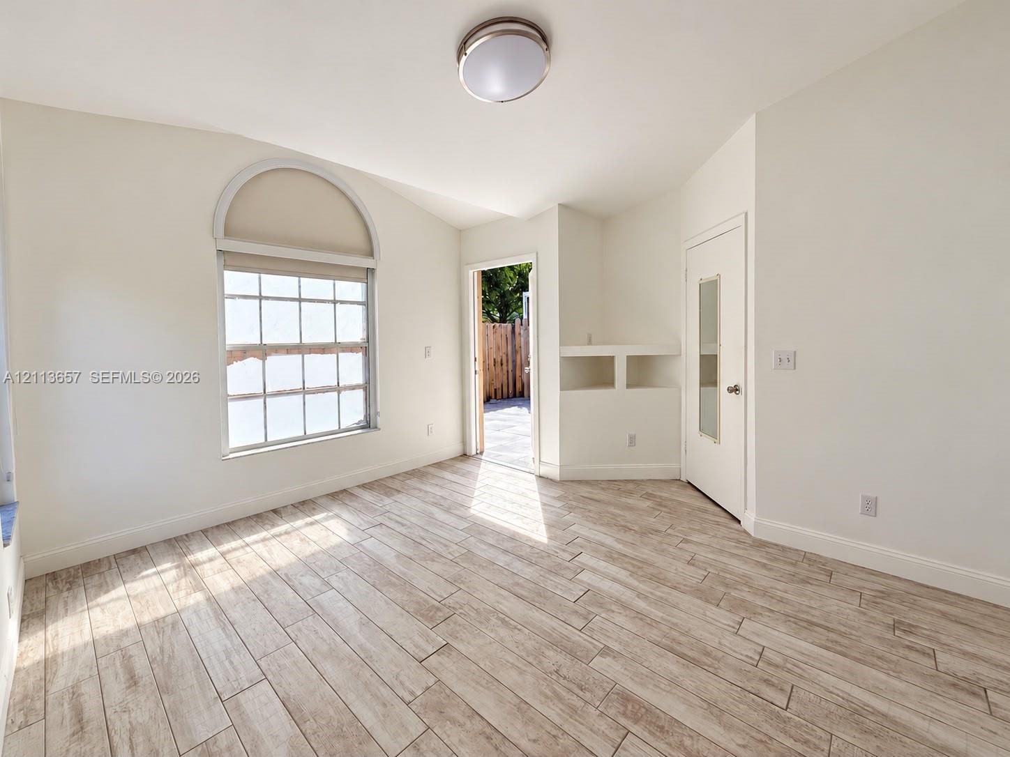 Room featuring wood-finish tile flooring, vaulted ceilings, a prominent arched window, built-in shelving, and a glass-paneled door Room featuring wood-finish tile flooring, vaulted ceilings, a prominent arched window, built-in shelving, and a glass-paneled door
