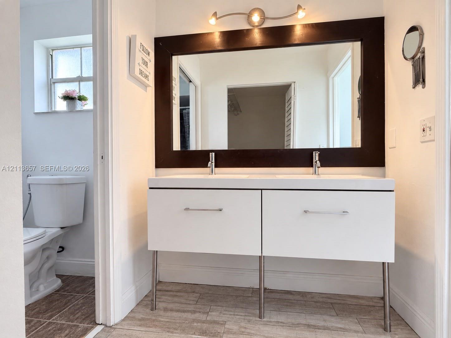Floating dual-sink vanity with matte white cabinetry, metal support legs, rectangular basin, and silver-tone hardware, featuring a large mirror with a dark wood-finish frame and overhead multi-bulb track lighting